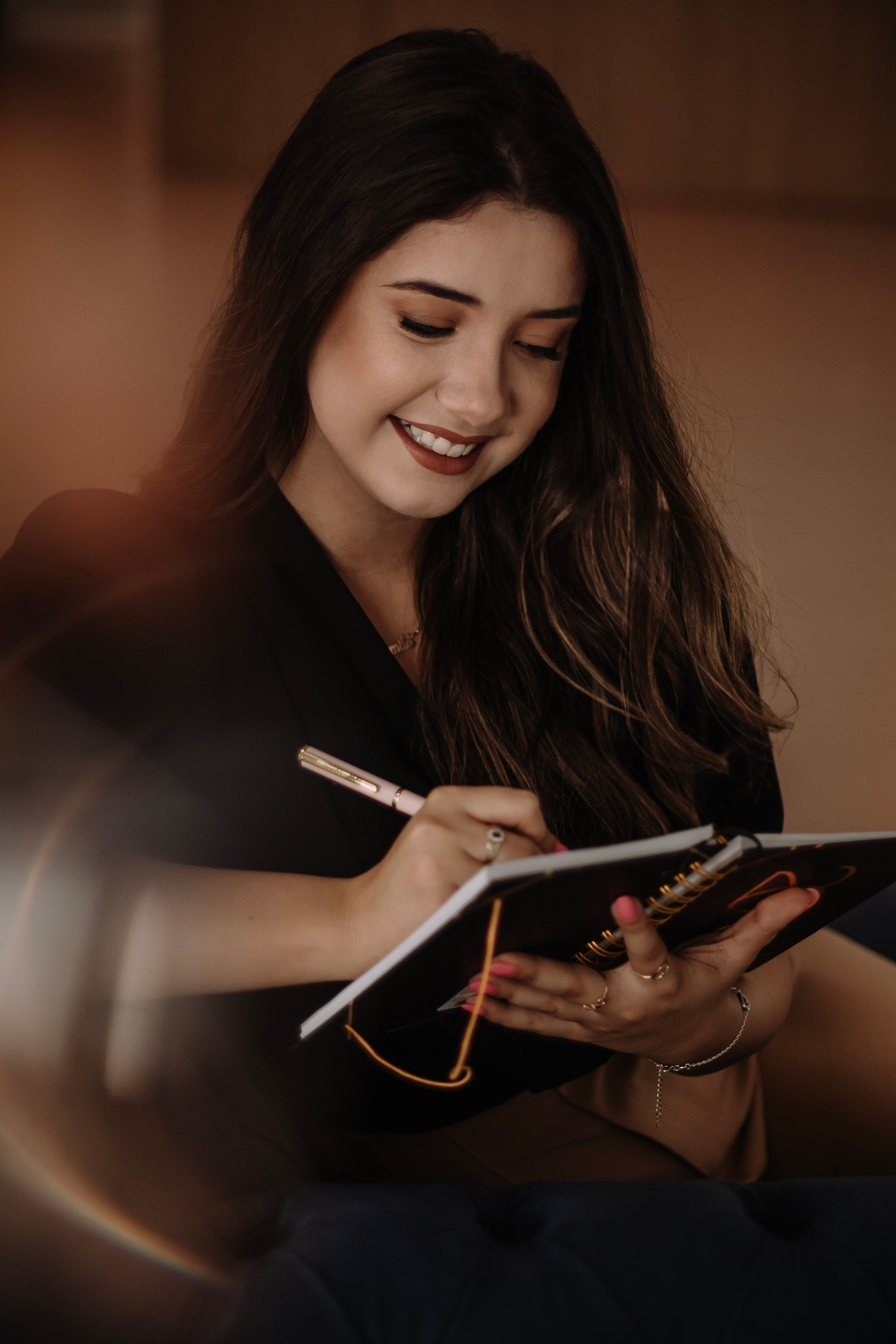 Psicóloga Kamila da Silveira. Uma mulher de cabelos longos, que está sentada em uma poltrona azul, sorrindo e fazendo anotações em sua caderneta.